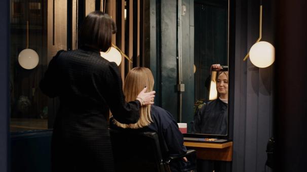 A woman is sitting in a salon looking at her new haircut with the hairdresser behind the chair.